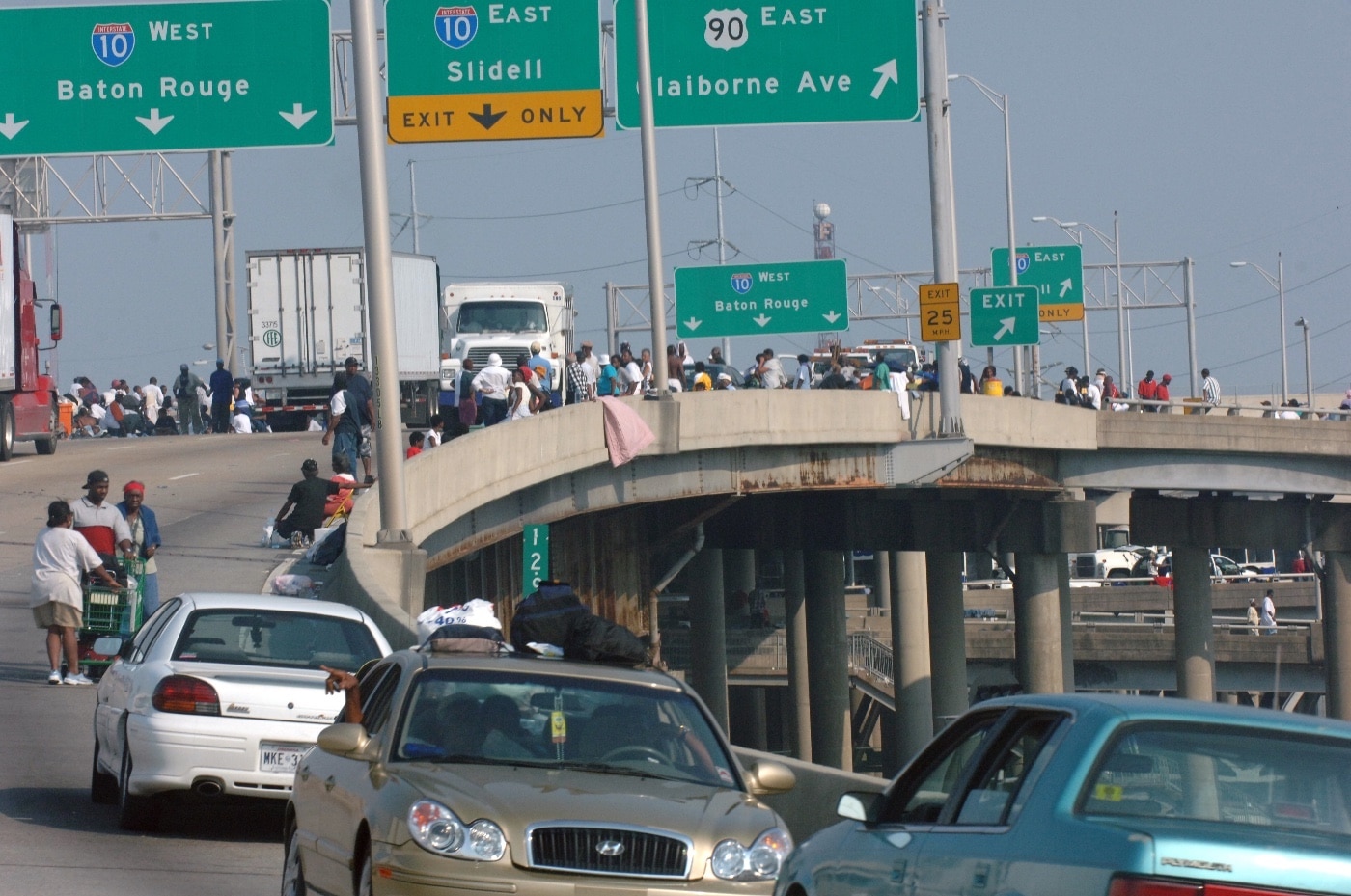 A large crowd of people gathered on an interstate bridge in New Orleans following Hurricane Katrina, their faces marked by frustration and desperation. Some individuals hold signs or gesture emphatically, while others sit or stand in clusters, shielding themselves from the sun. The scene reflects the scarcity of essential resources such as food and water in the aftermath of the hurricane, with no visible aid in sight. The damaged urban landscape in the background underscores the breakdown of infrastructure, including access to utilities and municipal water. This image captures the critical need for disaster preparedness, emergency response, and community support in urban areas affected by extreme weather events.