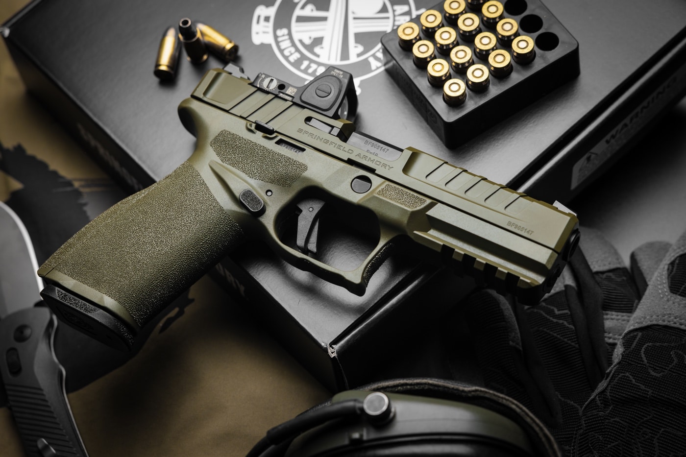 In this professional photograph, the OD Green version of the Springfield Echelon is prominent in the center of the image. Surrounding it are a number of other related items including 9mm ammo and electronic hearing protection.
