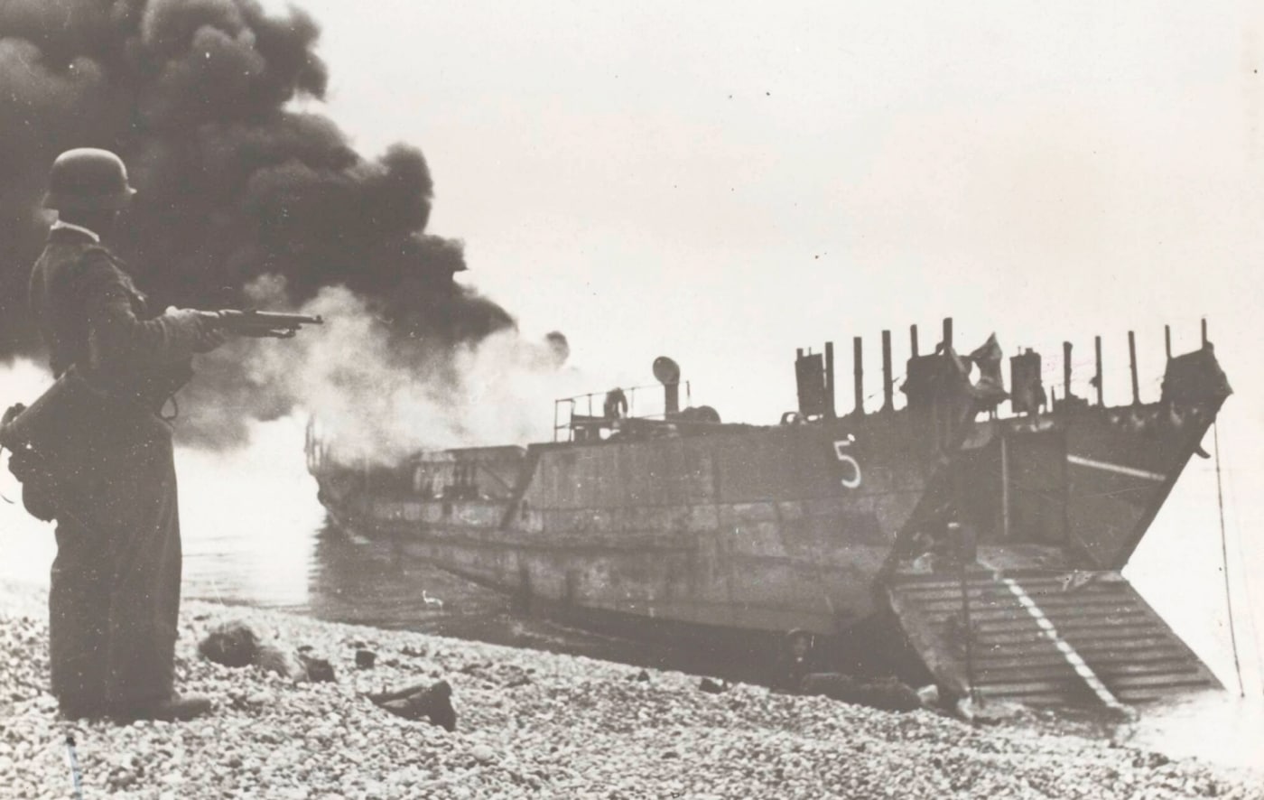 In this black and white photo from the Dieppe Raid, a German soldier with Kar 98k rifle stands in front of a burning British landing craft.