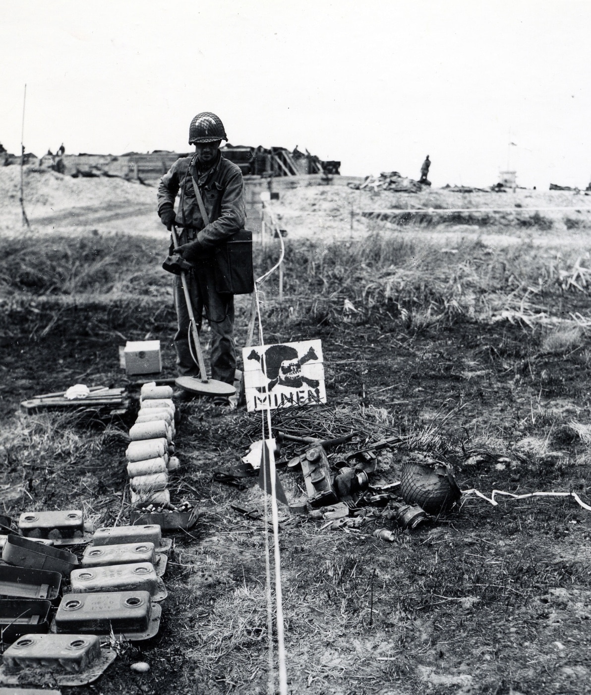 Here we see a U.S. soldier with a mine detector in Normandy at Atlantic Wall. Areas cleared were marked in white tape.