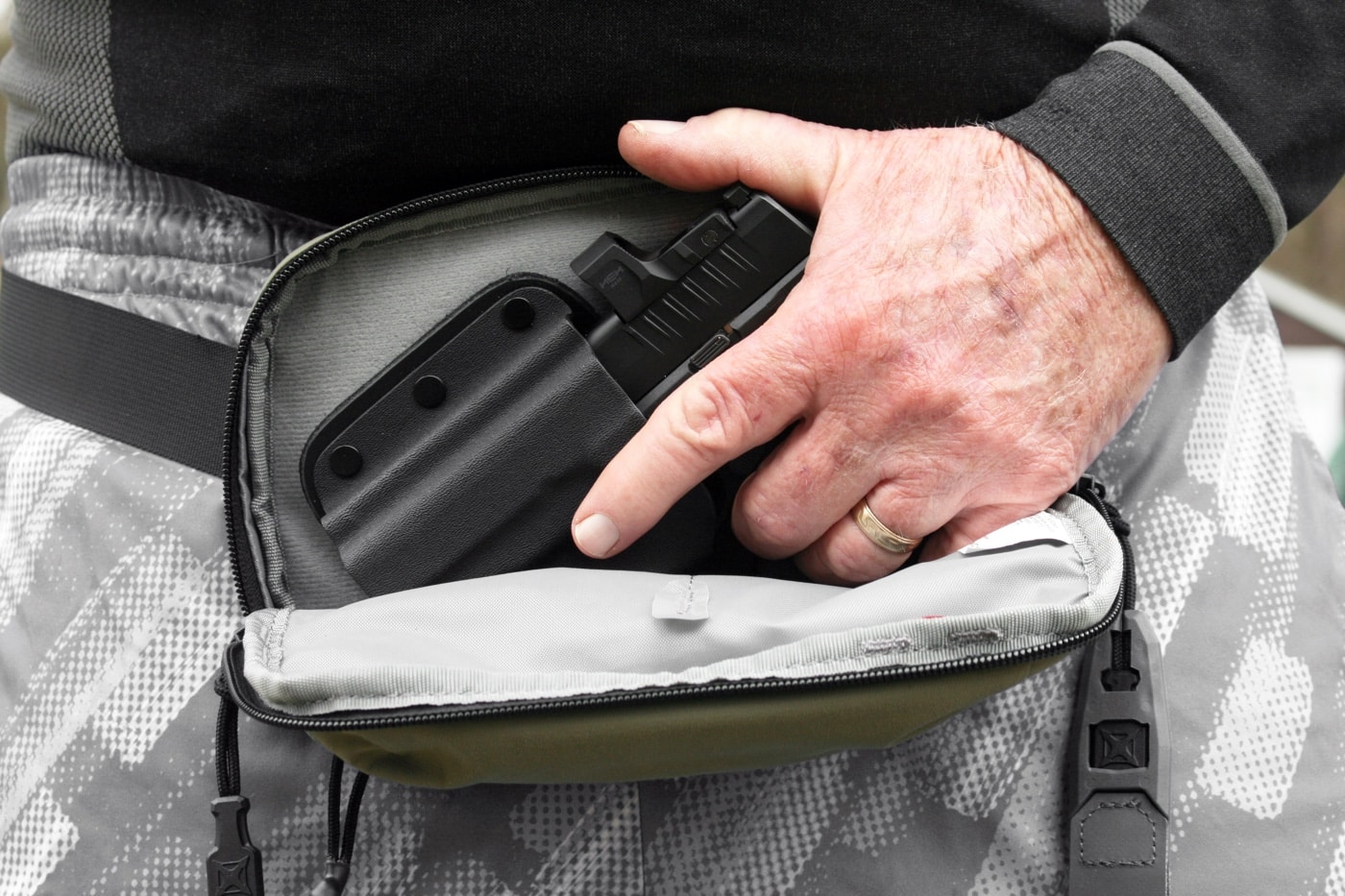 In this photo, we see the author practicing a draw of his Springfield Armory Hellcat — equipped with a red dot sight — from a unique carry bag. The bag uses Velcro and zippers to secure the handgun while not being used.