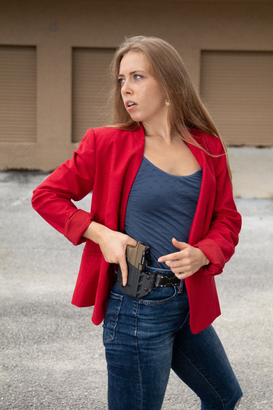 This photo is of a woman drawing a Springfield Hellcat OSP from a CCW holster. The woman is wearing denim blue jeans, a pullover blouse and a red suit jacket. She is also wearing a black leather belt. The strong side outside the waistband holster is made of Kydex.
