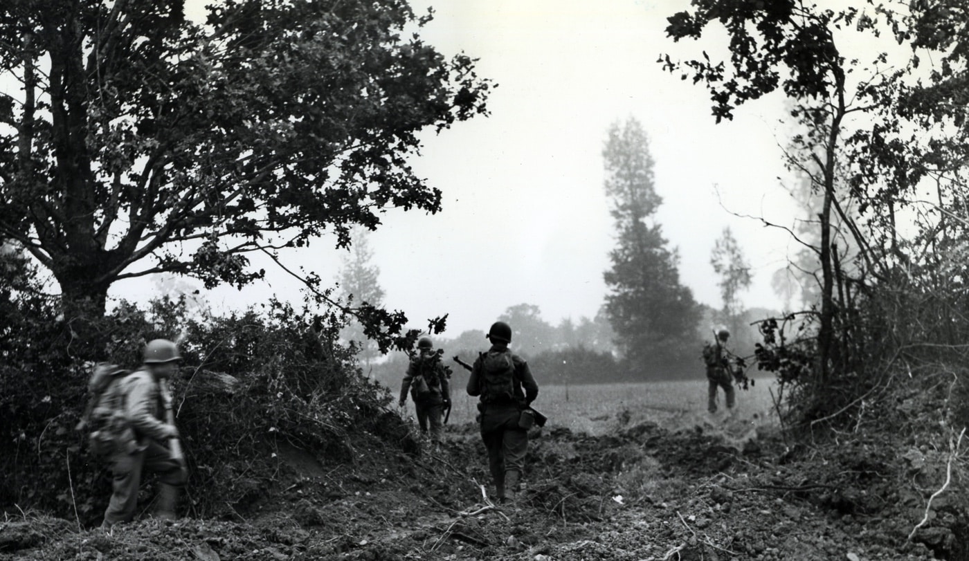 American GIs advance through a hedgerow opening in Normandy