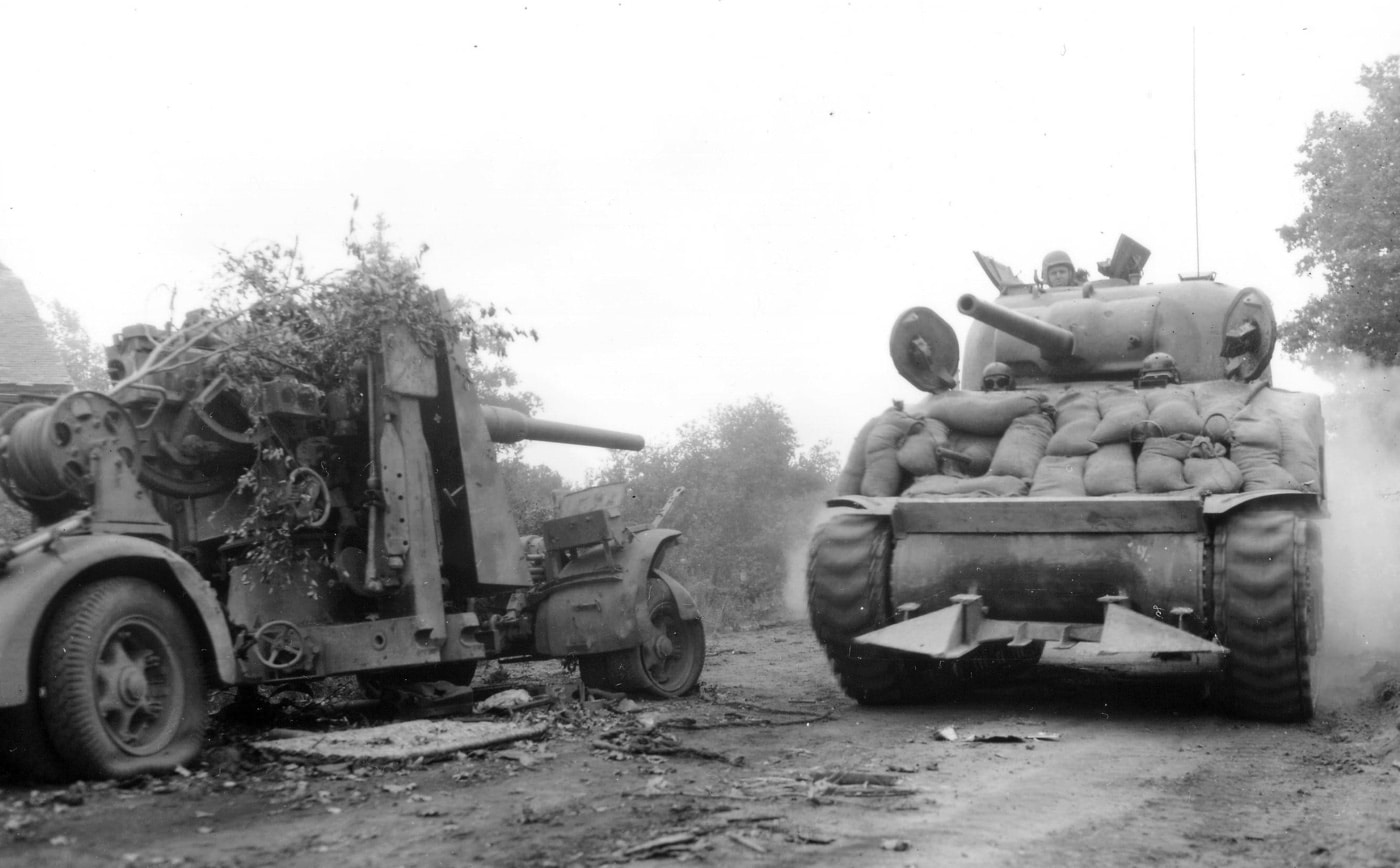 M4 Sherman tank with sandbag armor passes a destroyed German Flak 36 88mm