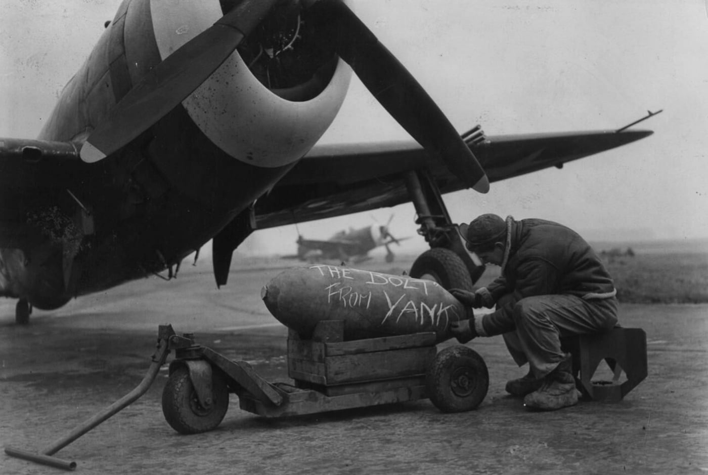 Sergeant Thomas J. Zettervall, from the 353rd Fighter Group, prepares a 500 lb bomb by inscribing "The bolt from Yanks" on its surface at Metfield Air Base. In the background, a P-47 Thunderbolt waits, symbolizing the adaptability of this aircraft to carry bombs as well as fight in the skies. The scene demonstrates the expanding role of the Thunderbolt from escort fighter to fighter-bomber. This image captures a moment of ingenuity and determination among Allied forces during World War II.