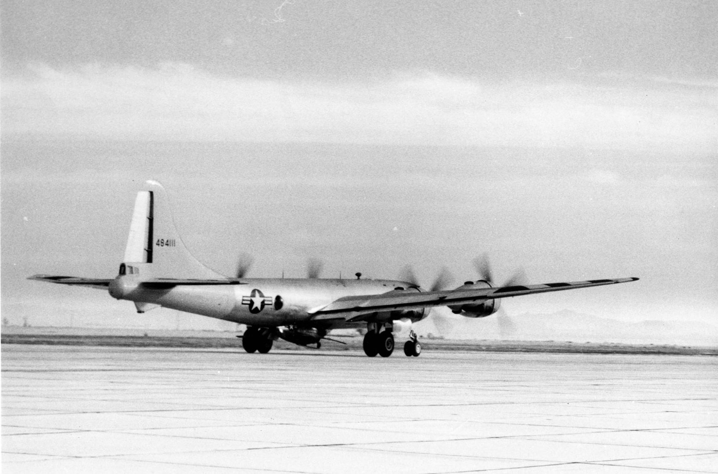 A B-29 Superfortress bomber is seen taking off with the McDonnell XF-85 Goblin attached underneath its fuselage. The compact Goblin, secured in a recessed position, was designed as a parasite fighter to defend bombers mid-mission. The image showcases the engineering challenges and innovative concepts tested in post-WWII aviation. The scene emphasizes the scale difference between the large bomber and the small, experimental jet.