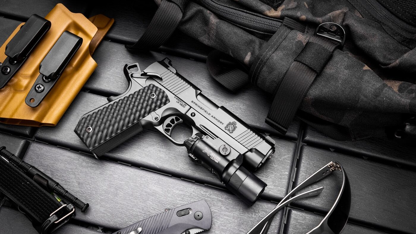 A Springfield Armory 1911 semi-automatic pistol with the Carry Contour cut is displayed on a table. The Carry Contour modification is visible, showcasing the bobbed rear grip frame that reduces printing when concealed. Surrounding the pistol are various pieces of shooting gear, including a holster and cleaning tools. The setup emphasizes the pistol's versatility as a concealed carry firearm while retaining its iconic design.