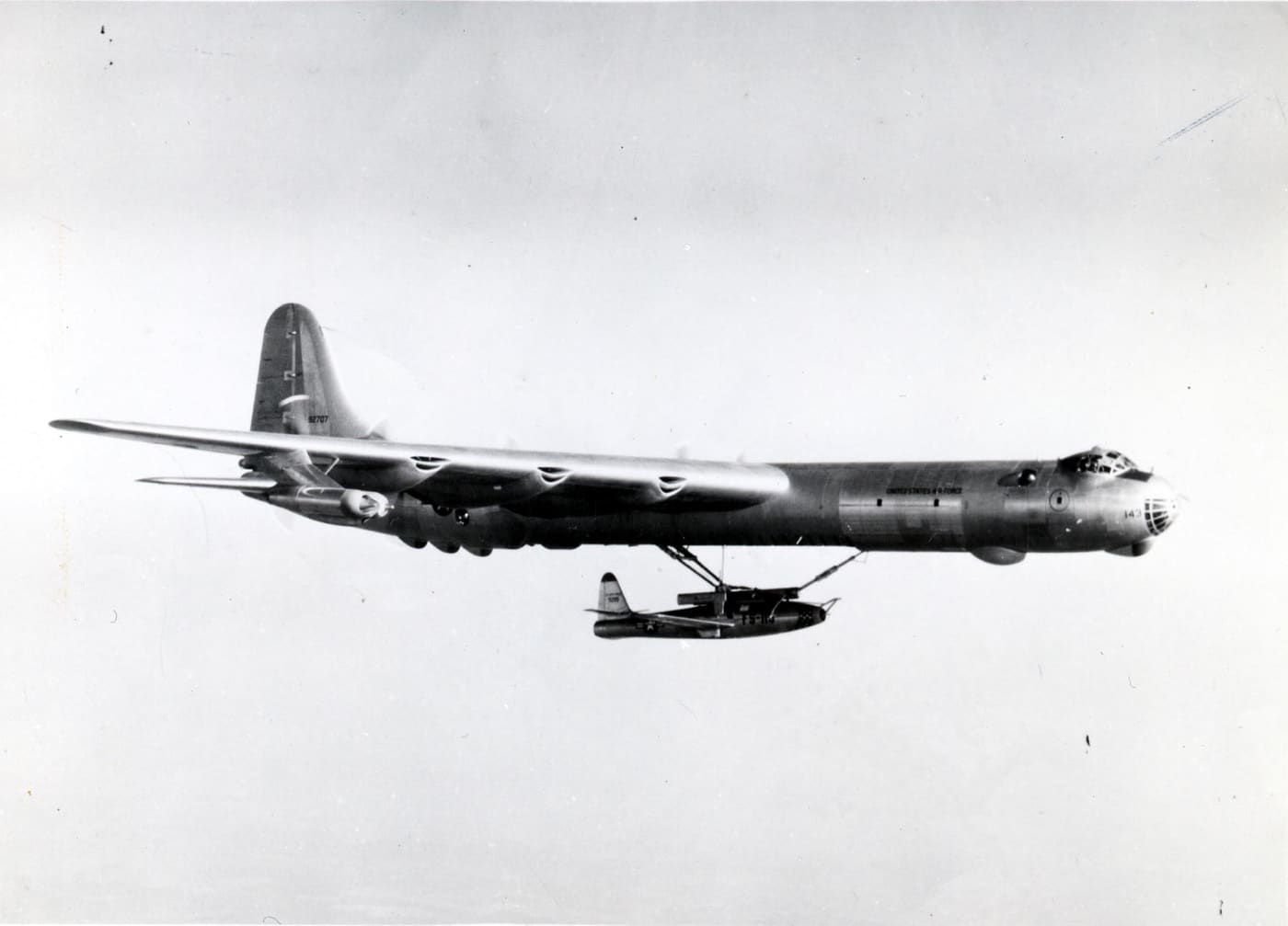 A Republic F-84E Thunderjet is captured in its extended position beneath a Convair B-36 Peacemaker during flight. The fighter jet hangs from a specialized cradle, designed to release and retrieve it mid-air as part of a parasite fighter program. The contrast between the small, sleek F-84E and the colossal B-36 bomber emphasizes the ambitious engineering of the era. This image showcases Cold War experimentation aimed at increasing bomber defense during long missions.