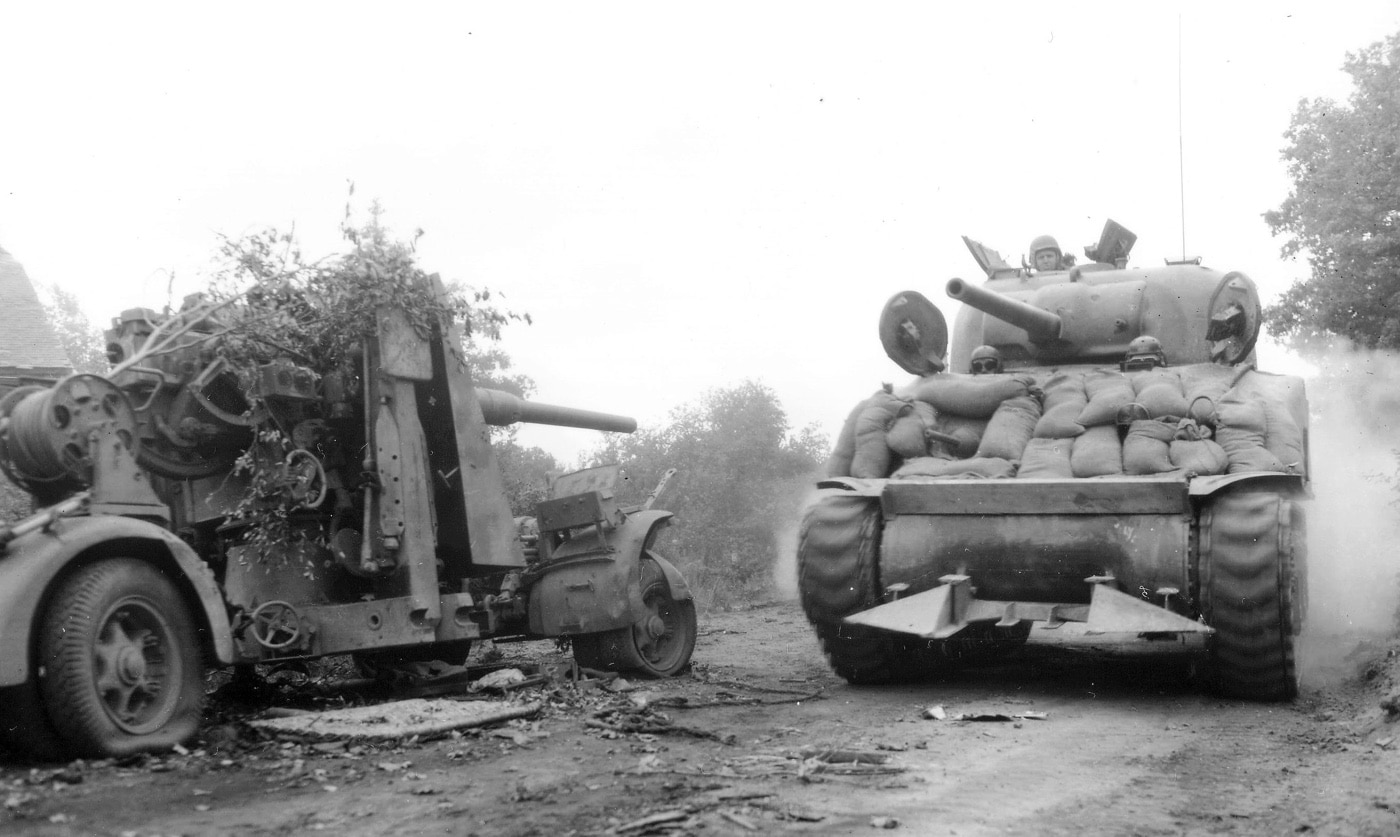 An M-4 Sherman Rhino tank advances through a Normandy field, passing a knocked-out German 88mm gun. The remnants of the 88mm, a feared weapon during the invasion, serve as a stark reminder of the intense fighting in the region. The tank’s hedgerow-cutting apparatus highlights the innovative strategies used by Allied forces to breach German defenses. This image symbolizes the shifting momentum of the war in favor of the Allies.