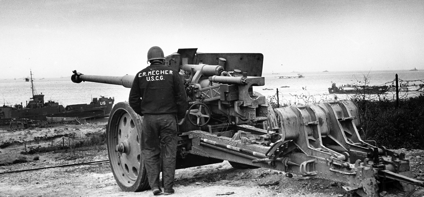 A U.S. Coast Guard sailor stands beside a captured German Pak 43/41 anti-tank gun at Normandy during Operation Neptune in 1944. The massive gun, known for its devastating firepower, was a significant threat to Allied forces during the invasion. Its presence here symbolizes the Allies’ success in overcoming German defenses along the beaches. The sailor’s stance next to the weapon reflects a moment of triumph amidst the challenges of the Normandy campaign.