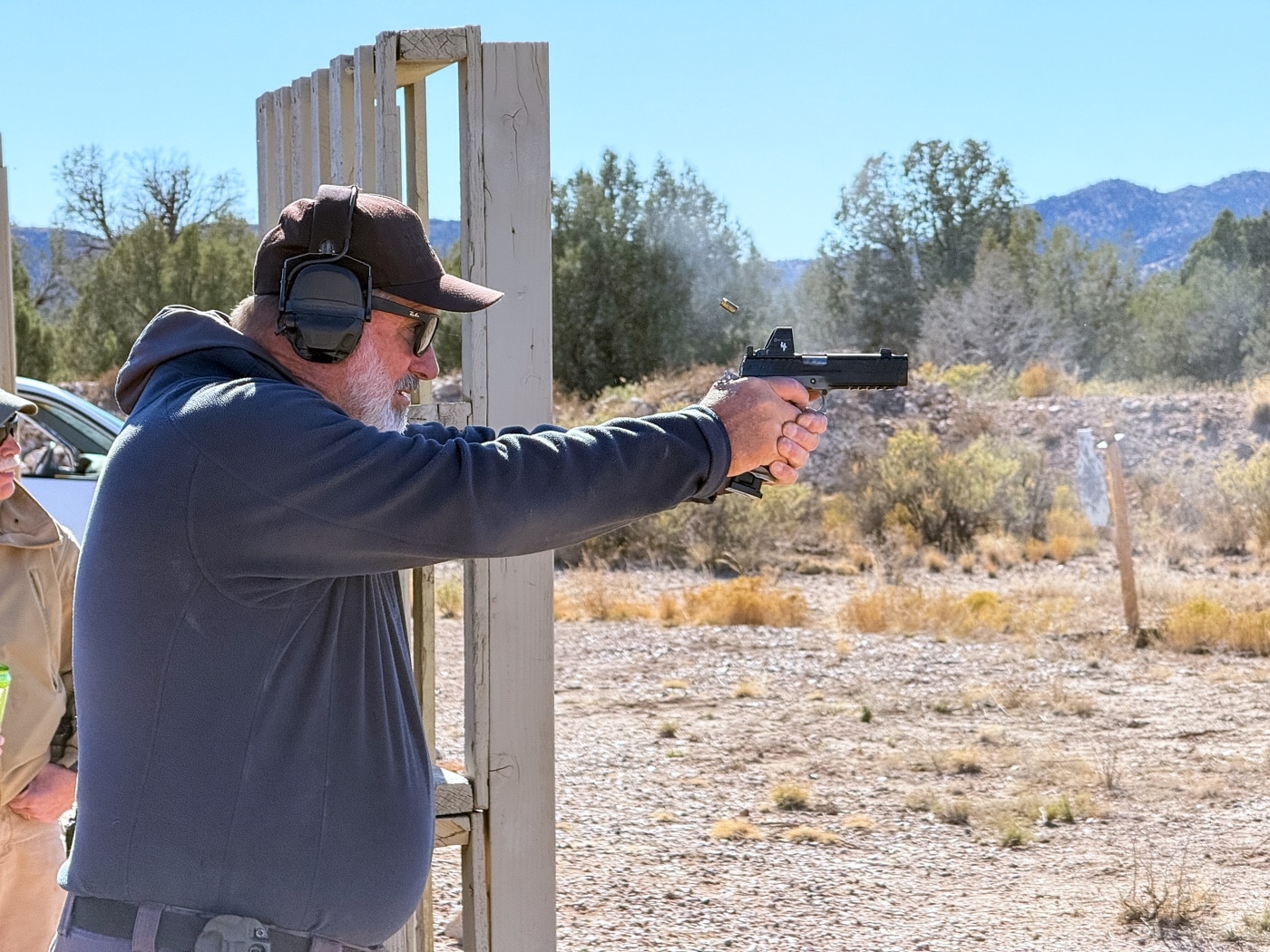 Rob Leatham is pictured on the shooting range at Gunsite Academy, expertly demonstrating proper shooting techniques with a Springfield Armory pistol. His posture reflects a solid shooting stance, with a firm grip on the firearm and full focus on the target. The pistol’s sleek design is prominently visible as he fires a shot. The range is surrounded by rugged desert terrain, and steel targets are placed in the background. This photo captures a moment of precision and skill, illustrating Rob’s teaching prowess and his emphasis on accuracy and control.