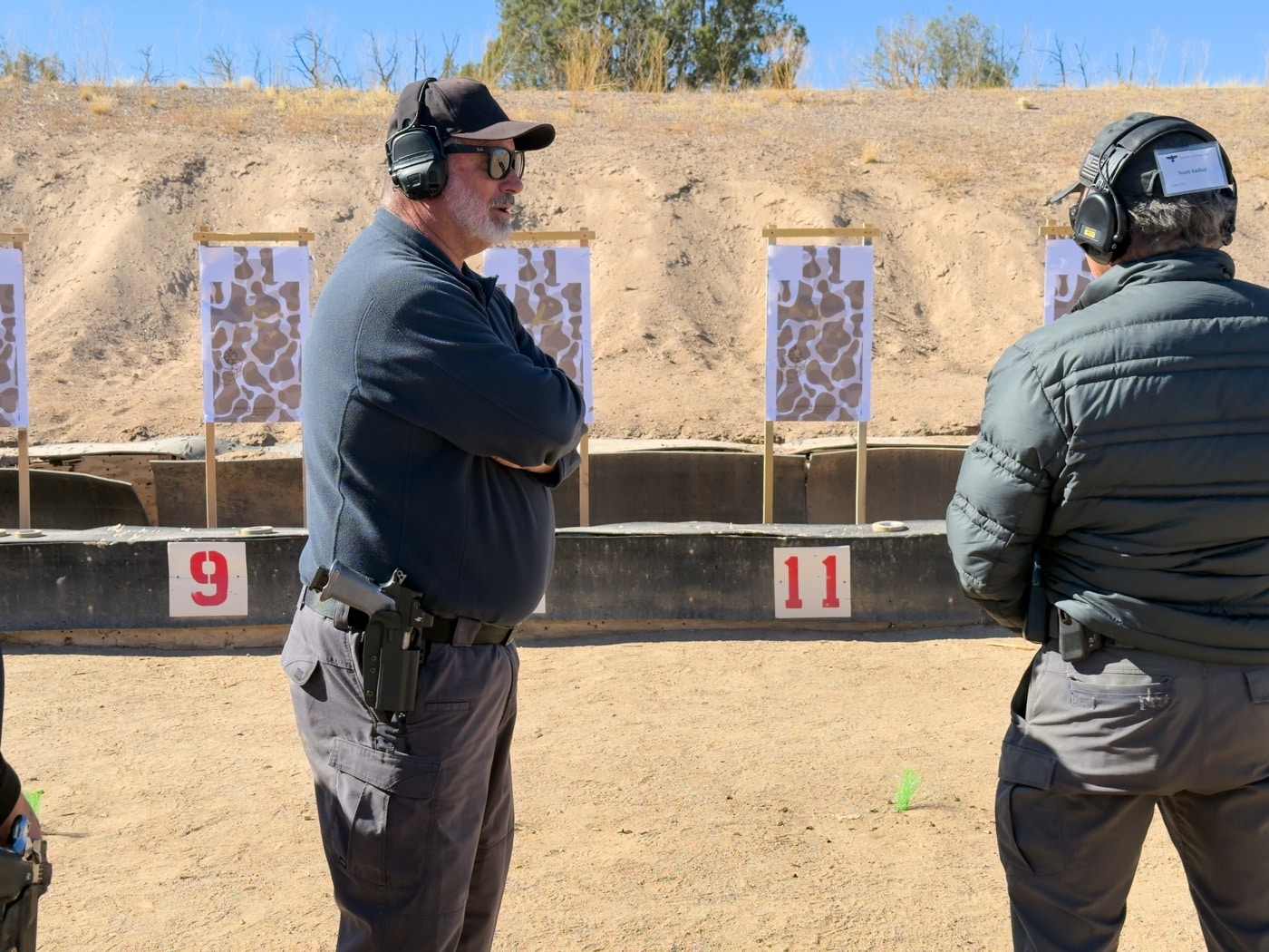 Rob Leatham, an experienced professional shooter and instructor, is pictured providing one-on-one coaching to a student on the Gunsite Academy shooting range. The student stands in a firing stance, gripping a pistol, while Rob observes closely, offering detailed feedback and guidance. The interaction demonstrates Rob’s hands-on teaching approach, focusing on improving accuracy, speed, and technique. The outdoor range features sandy terrain and steel targets in the background, creating a practical and focused training environment. This image highlights the value of personalized instruction in developing advanced shooting skills.