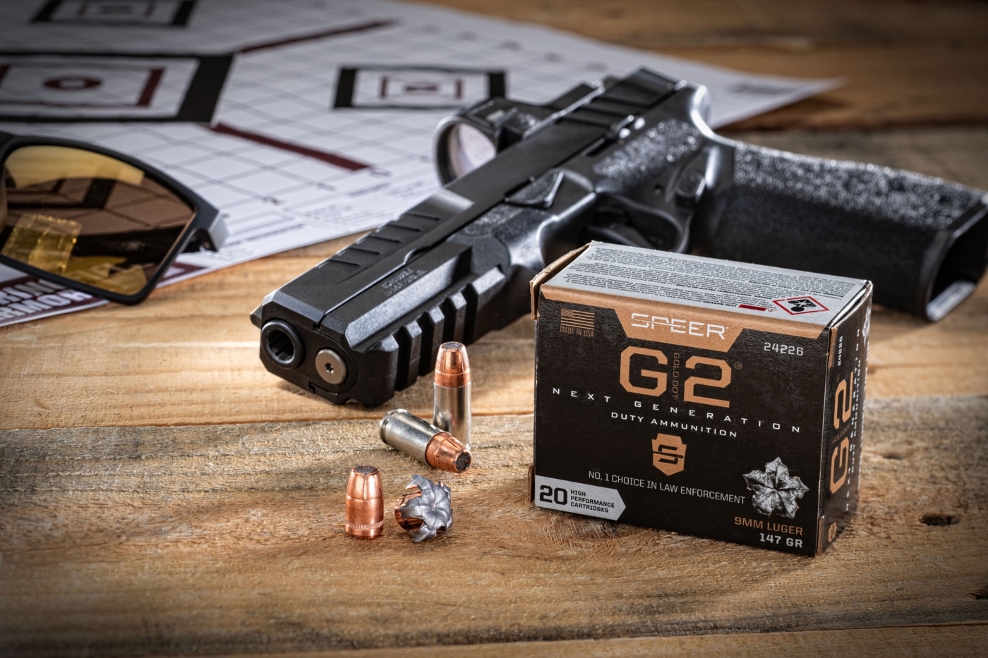 A photograph showing Speer Gold Dot G2 9mm Parabellum ammunition prominently displayed in the foreground. The brass-cased cartridges have a clean, polished appearance with nickel-plated casings and hollow points, indicative of their premium defensive design. In the background, a Springfield Armory Echelon handgun rests on the table, positioned for visibility but slightly out of focus. Behind the firearm, paper targets with visible bullet holes are faintly visible, indicating a range test scenario. The composition highlights the ammunition's performance and reliability, combined with a modern, high-performance handgun. This setup is ideal for showcasing defensive shooting and ammunition testing results.