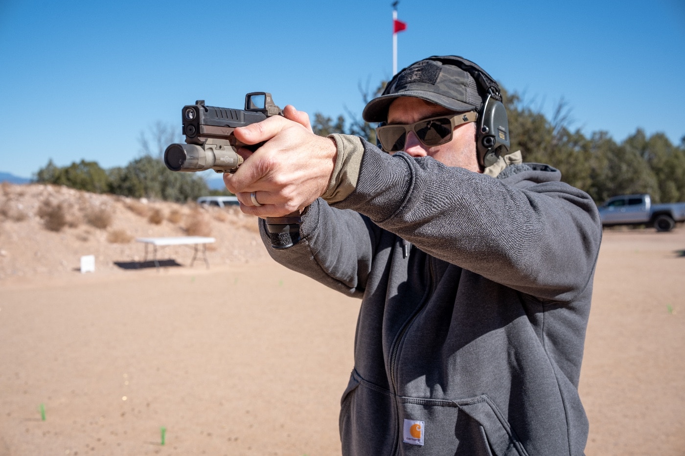 The author is captured on the Gunsite Academy shooting range, firing a Springfield Armory Echelon pistol as part of an advanced training class. Their shooting stance is firm and focused, with both hands gripping the pistol and their eyes locked on the target. The black-and-gray Echelon pistol stands out against the natural outdoor backdrop of the range, which features steel targets and rugged desert terrain. This image conveys the intensity and practical application of the skills being developed during the class, emphasizing the importance of accuracy and proper technique.