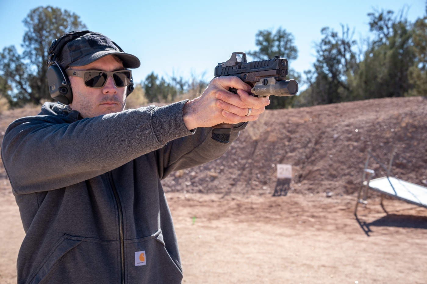 The author is shown on the Gunsite Academy range firing a Springfield Armory Echelon pistol, equipped with custom accessories detailed in the article. The A.C.E. grip module from Icarus Precision features aggressive texture and ergonomic enhancements for improved handling. A PRP trigger kit ensures a lighter, more responsive pull, while the EOTech EFLX Mini Red Dot optic provides a clear and expansive field of view for fast target acquisition. The author maintains a steady shooting stance, illustrating the benefits of the performance upgrades. The surrounding desert terrain and steel targets underscore the practical and challenging training environment.