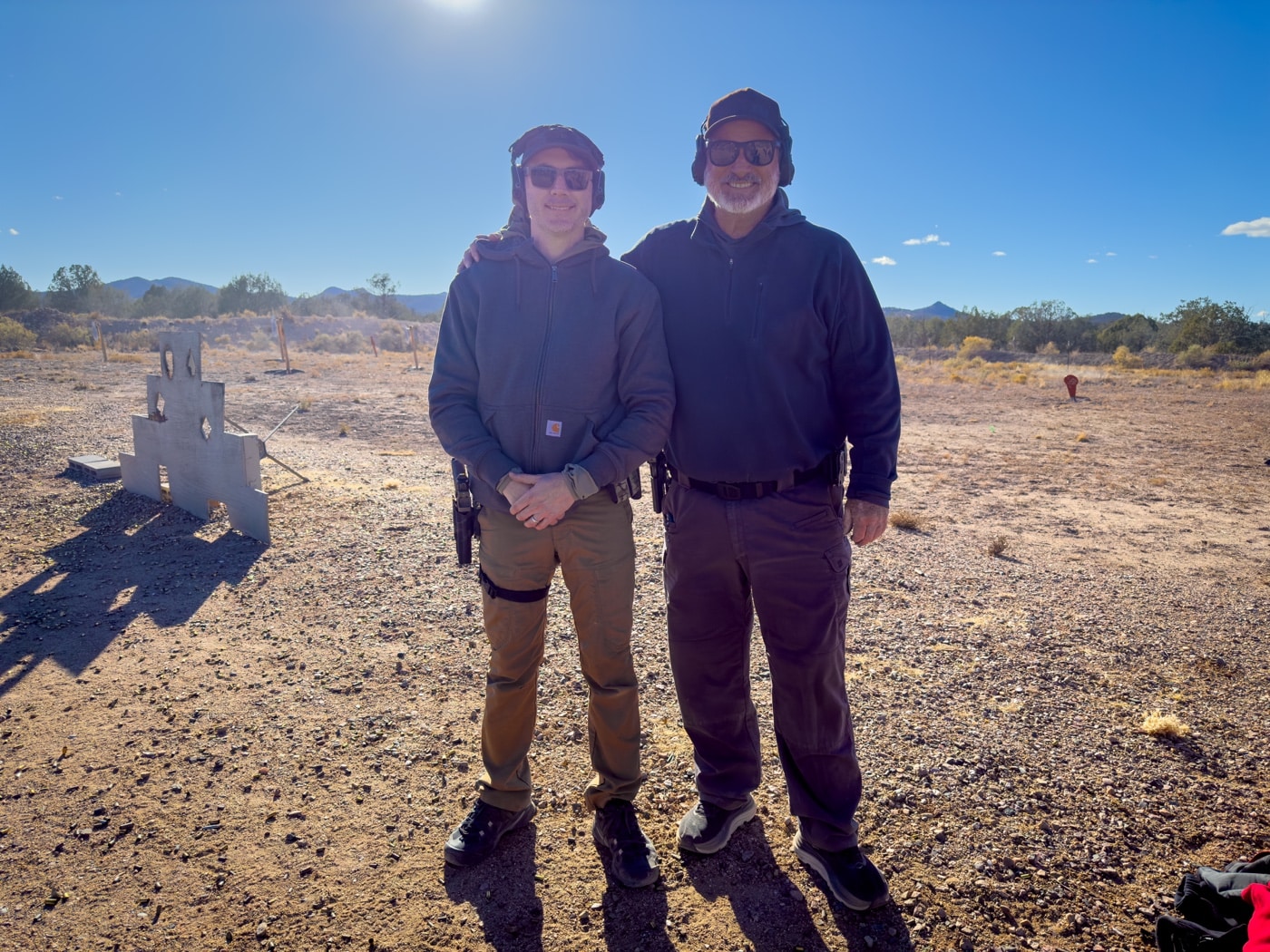 The author stands beside Rob Leatham, a renowned professional shooter and instructor, at the end of the Performance Pistol Symposium at Gunsite Academy. Both are smiling, reflecting the camaraderie and accomplishments achieved during the class. The author holds his Springfield Armory Echelon pistol, highlighting the tools and techniques practiced throughout the training. The setting includes the outdoor shooting range with desert terrain in the background, symbolizing the practical and immersive environment of the course. This photo captures the mutual respect and positive atmosphere of the advanced training experience.