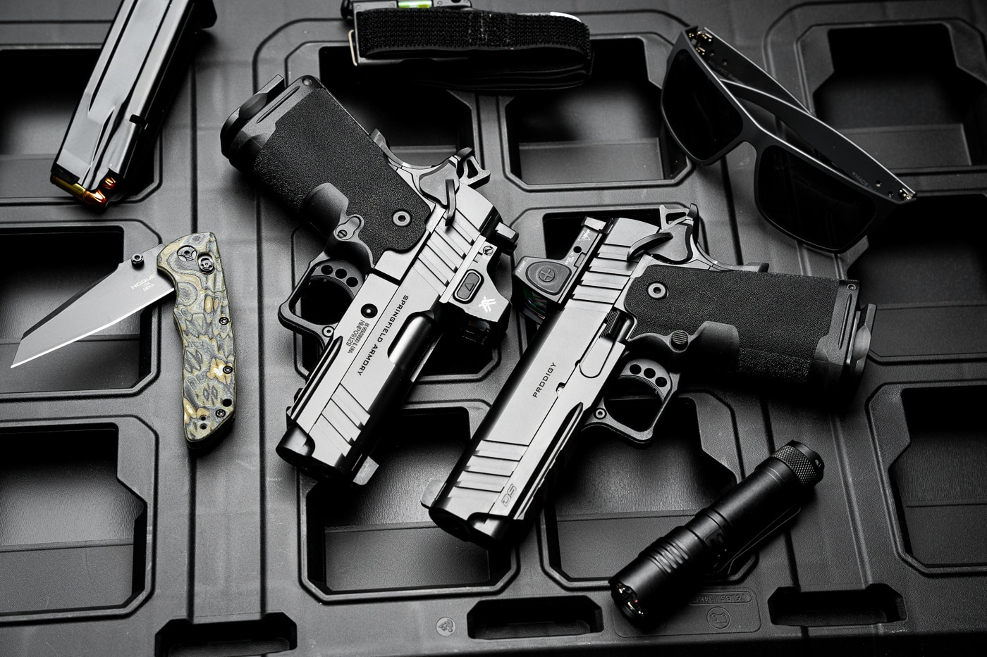 A lifestyle photo featuring two Springfield Armory 1911 DS Prodigy Compact pistols, one with a 3.5-inch barrel and the other with a 4.25-inch barrel. Surrounding the pistols are common everyday carry (EDC) items, including a pocket knife, a compact flashlight, a pair of sunglasses, and a spare magazine. The pistols' matte finish and compact designs are emphasized, making them ideal for concealed carry. The arrangement suggests practicality and readiness for defensive scenarios.