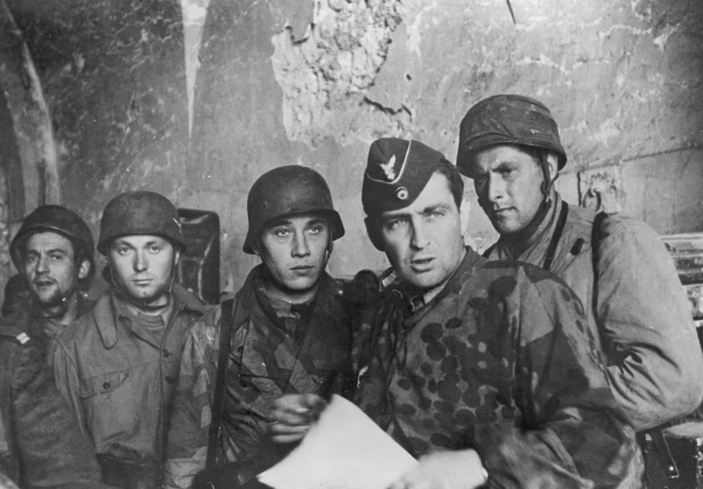 A group of German Fallschirmjäger paratroopers from the 1st Parachute Corps, known as the Green Devils, during the Battle of Monte Cassino in 1944. The soldiers are shown wearing distinctive camouflage smocks and helmets, which were characteristic of their elite status within the German military. They are positioned amidst rocky terrain, indicative of the rugged environment surrounding Monte Cassino and the Gustav Line. These paratroopers played a crucial role in defending the fortified positions against relentless Allied assaults. The image highlights the tenacity and preparedness of German forces during one of the most fiercely contested battles of the Italian Campaign. The Fallschirmjäger's presence underscores the difficulty the Allies faced in overcoming well-trained and entrenched defenders.