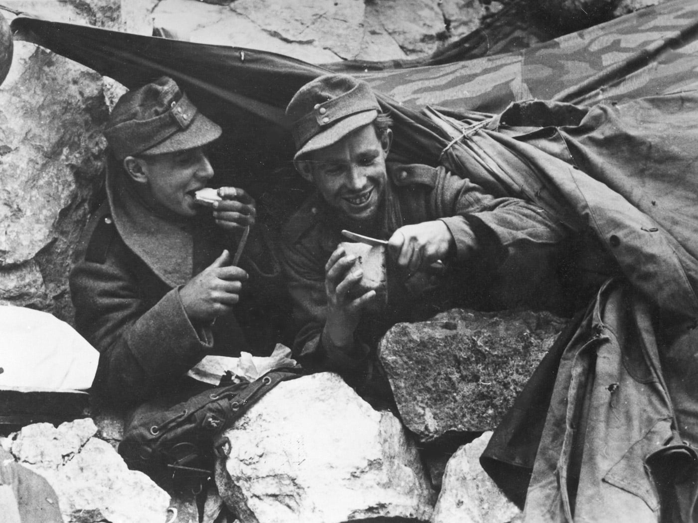 A primitive hut constructed between rocks by German soldiers during the Battle of Monte Cassino, providing basic shelter on the front lines. Two soldiers are visible in the scene, seated and eating a meal amidst the rugged terrain. The makeshift structure reflects the harsh conditions faced by both sides during the prolonged battle for control of the Gustav Line. The rocky surroundings emphasize the challenging environment of Monte Cassino, which played a significant role in the defensive strategies of the German forces. This image captures a moment of respite amidst the intense combat of the Italian Campaign.