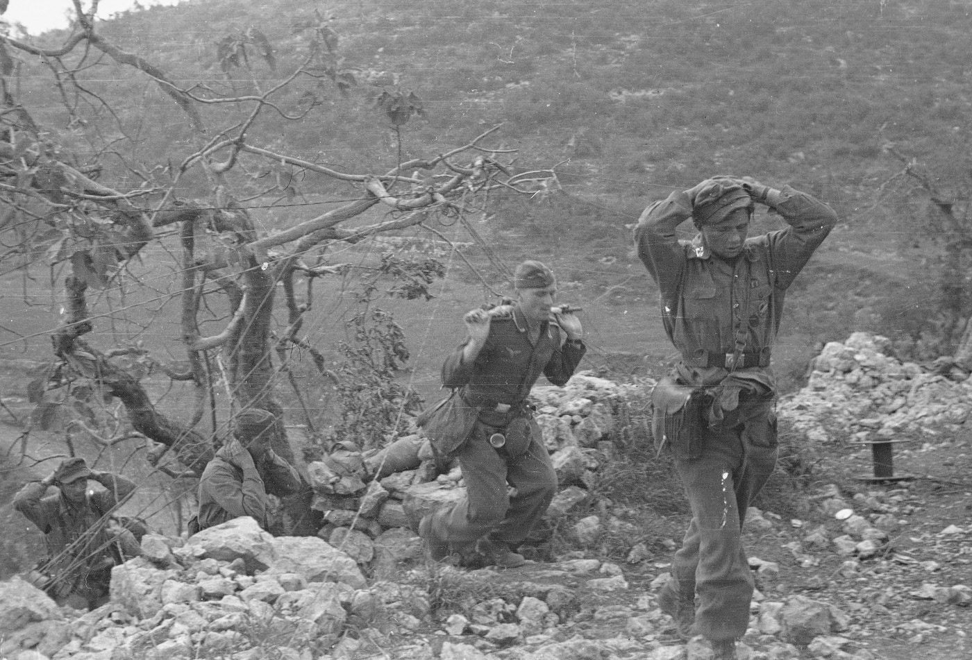 A group of German prisoners walks along a narrow mountain path on their way to captivity during the Battle of Monte Cassino. The soldier on the right is identifiable by a map case strapped to his side and a visible parachutist's badge, indicating he is a member of the elite Fallschirmjäger paratroopers. The rugged terrain emphasizes the challenges of the Italian Campaign, where mountainous landscapes played a significant role in the defensive strategies of the German forces. The prisoners appear weary, reflecting the intense combat that led to their capture during the Allied push to break through the Gustav Line. This image captures a moment of transition as the tide of battle shifts in favor of the Allies.