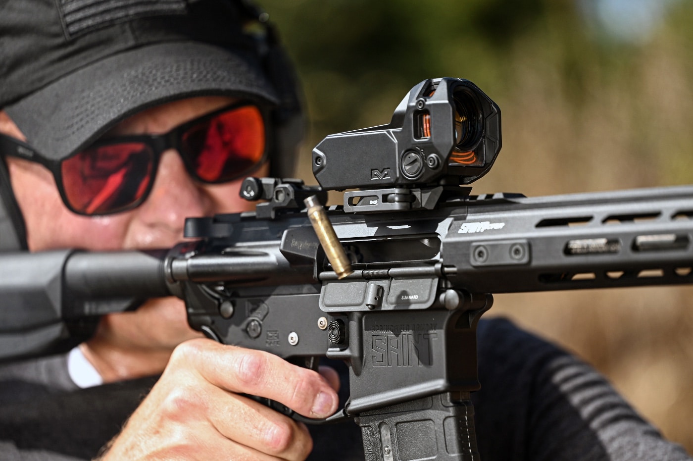 A closely cropped image shows the author firing a Springfield Armory SAINT rifle on a shooting range, equipped with the Meprolight M22 red dot sight. Bright daylight highlights the optic's reticle, powered by fiber optic and tritium technology. Spent 5.56 NATO brass is seen being ejected from the rifle, emphasizing the firearm's live fire test. The sight's compact and battery-free design is evident, making it a reliable option for dynamic shooting conditions. Dust and movement add to the realism of the shooting environment, showcasing the optic's practical use.