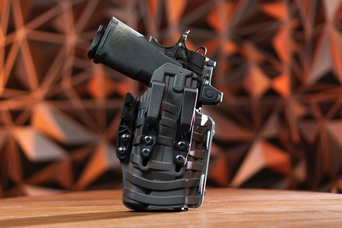 A PHLster Floodlight2 holster stands upright on a table with a Springfield Armory 1911 DS Prodigy pistol securely inserted. The holster's sleek design and ComfortScape texture are visible, showcasing its focus on comfort and concealment. The bright red bungee cord, used for retention adjustment, adds a pop of color to the otherwise black holster. This universal holster accommodates various full-size and compact handguns equipped with weapon-mounted lights, such as the SureFire X300. The open-ended design supports accessories like suppressor-height sights and threaded barrels. The pistol’s ergonomic grip and the holster's discreet clips hint at the pair's concealment effectiveness. Ideal for concealed carry enthusiasts, this setup exemplifies a balance of innovation and practicality.