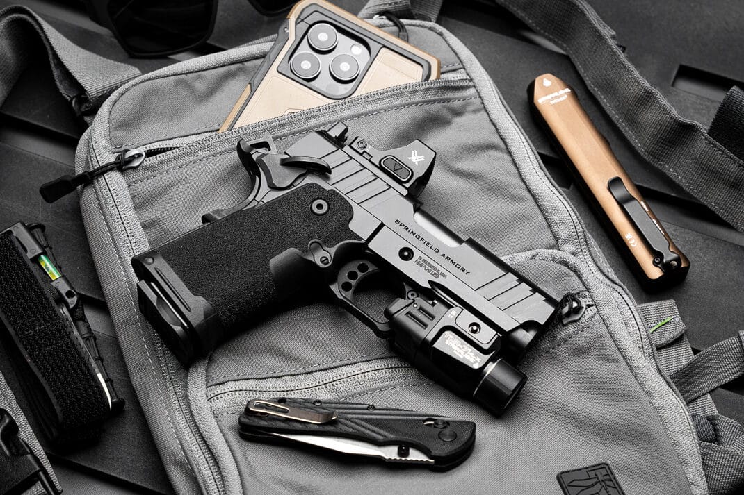 A Springfield Armory 1911 DS Prodigy Compact pistol with a 3.5-inch barrel is displayed lying on top of a backpack. The pistol is outfitted with a Vortex red dot sight for precision aiming and a Streamlight TLR-7 weapon-mounted light for low-light capability. The gun’s matte finish contrasts with the backpack’s textured fabric, emphasizing its compact and tactical design. The setup highlights its suitability for everyday carry or defensive applications in various environments.