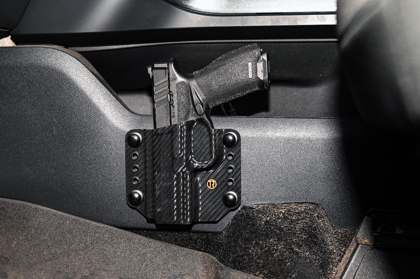 A Springfield Armory Echelon pistol sits securely in a custom holster mounted inside a truck, positioned for easy access. The holster is attached to the side of the center console, ensuring the firearm is both secure and readily available. The pistol's ergonomic design and sleek black finish are visible, reflecting its modern appeal and reliability. The truck's interior shows a clean, organized layout, emphasizing the practical setup for self-defense readiness. The scene is brightly lit, illustrating how a well-placed holster can help maintain situational awareness during emergencies. This configuration highlights the importance of responsible firearm storage within a vehicle. It demonstrates the balance between safety, accessibility, and preparedness.