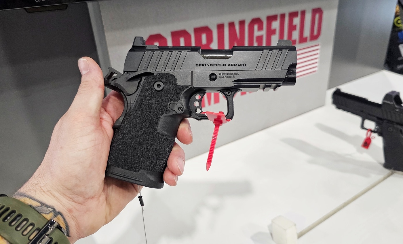 The author holding a Springfield Armory 1911 DS Prodigy Compact pistol inside the company’s exhibit booth at the 2025 SHOT Show. The firearm has a matte black finish and a contoured grip, making it a standout in the booth’s display. Behind him, a brightly illuminated backdrop showcases Springfield Armory’s brand logo and a variety of firearms, including rifles and handguns. The bustling environment captures the energy of the event, with other attendees inspecting displays and engaging with Springfield’s staff. The compact design and advanced features of the Prodigy Compact emphasize its appeal as a modern concealed carry option for visitors exploring the booth.