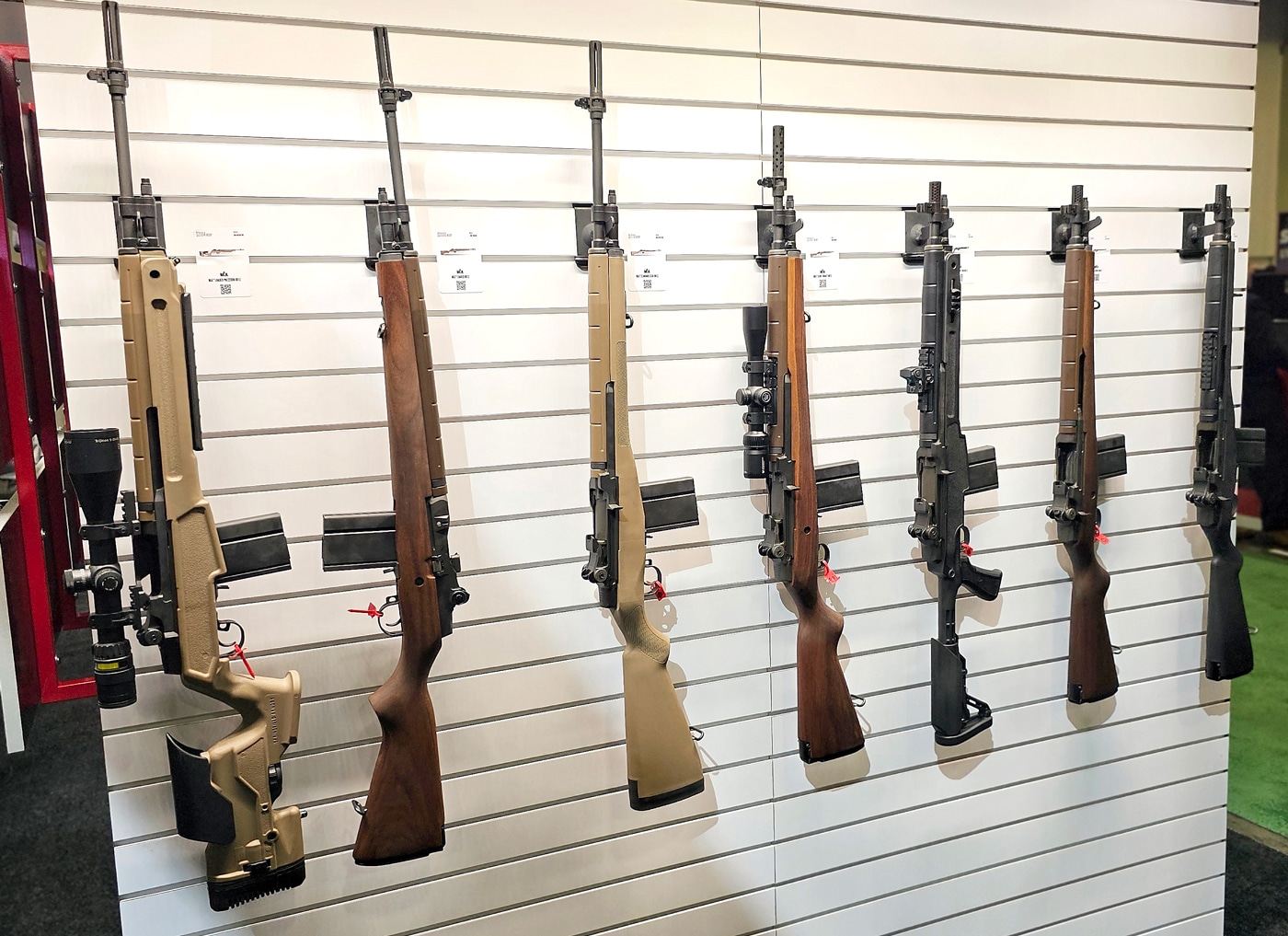 A display row featuring multiple Springfield Armory M1A rifles at the 2025 SHOT Show. The rifles are arranged in a clean, orderly line, with each model showcasing unique configurations, including the SOCOM 16 with its compact design and the Loaded Precision with its extended barrel and adjustable stock. The SOCOM 16 has a distinctive muzzle brake and a scout-style Picatinny rail, while the Loaded Precision boasts match-grade sights and a long-slotted flash suppressor. Visitors at the booth can be seen in the background, engaging with staff and examining other displays. The lighting emphasizes the rifles’ finishes, drawing attention to Springfield’s craftsmanship and attention to detail. This display highlights the versatility and range of the M1A lineup, appealing to both tactical and precision shooting enthusiasts.