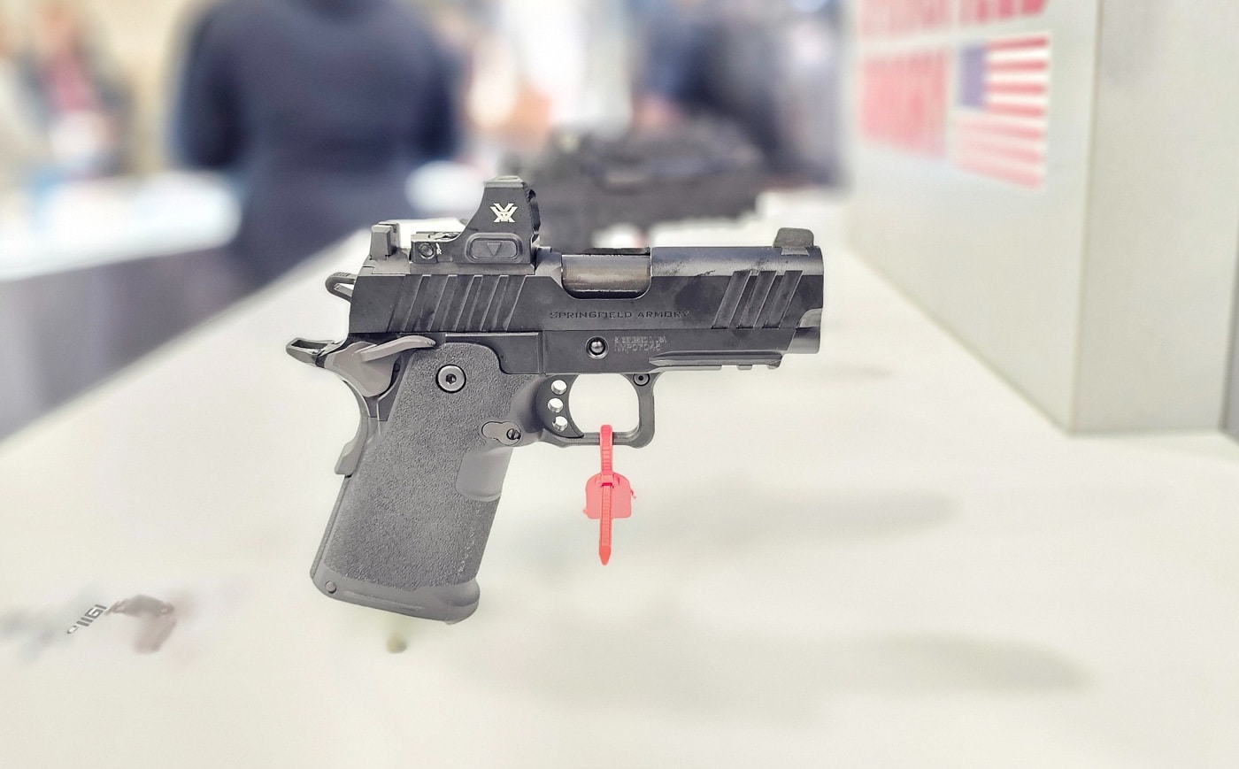 The Springfield Armory 1911 DS Prodigy Compact sits elegantly on a display stand at the 2025 SHOT Show, highlighting its compact design tailored for concealed carry. The 3.5-inch stainless steel bull barrel is finished with a black DLC coating, giving it a sleek and durable appearance. The shortened grip module and re-contoured frame enhance concealability while maintaining the ergonomic comfort Springfield is known for. Subtle front slide serrations add a functional aesthetic, complementing the optics-ready slide equipped with the Agency Optic System (AOS). Bright lighting emphasizes the pistol’s premium finish and modern design, drawing the attention of nearby visitors in the bustling booth. The Prodigy Compact is accompanied by two 15-round magazines, offering a blend of compact size and ample firepower. This display highlights Springfield’s commitment to creating innovative firearms for both personal defense and professional use.