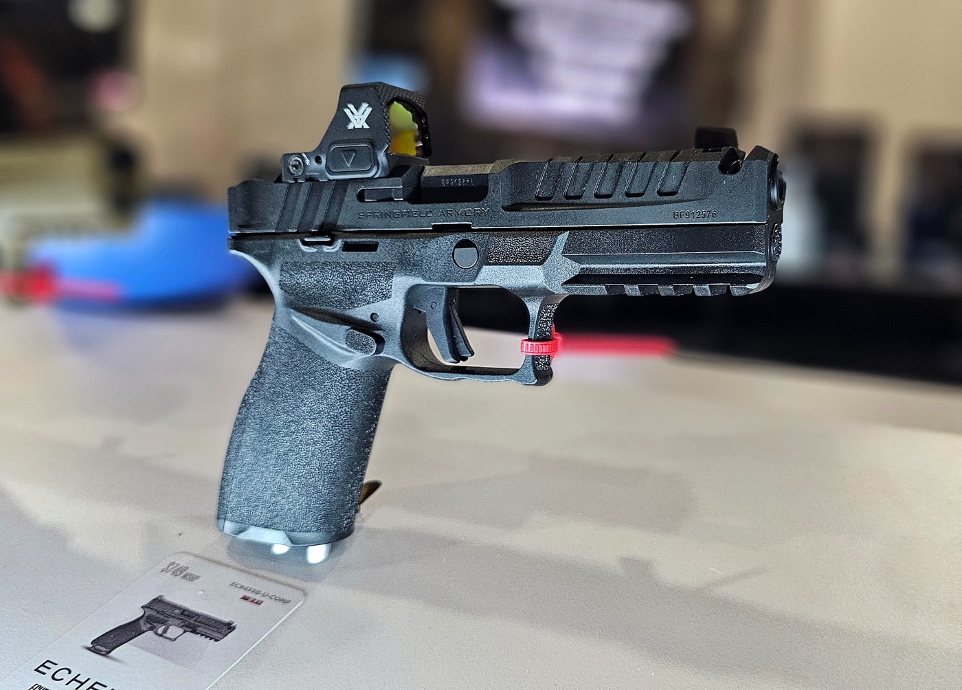 The Springfield Armory Echelon Comp pistol is displayed on a sleek pedestal at the 2025 SHOT Show booth. The 4.5-inch barrel features a discreet integrated compensator designed to reduce muzzle rise and enhance shooting precision. The pistol’s matte finish and clean lines are complemented by the variable optics interface system, making it compatible with a wide range of optics. Attendees in the background can be seen observing other firearm displays, adding to the vibrant and professional atmosphere of the booth. The Echelon Comp is presented with its accompanying 15-round magazines, emphasizing its versatility for both tactical and personal defense use. Bright lighting highlights the pistol's modern aesthetic and Springfield’s attention to detail in design and engineering. This display showcases Springfield Armory's commitment to innovation in the competitive handgun market.