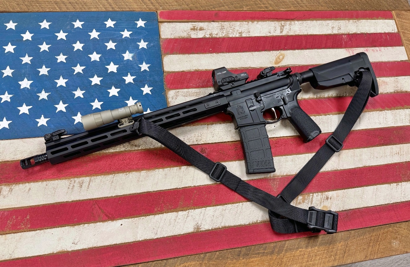 A SureFire Mini Scout Light Pro flashlight is attached to the M-LOK rail of a Springfield Armory SAINT rifle, resting on a wooden American flag. The flashlight’s low-profile mount ensures a snug fit along the rifle's foregrip. The rifle’s black finish contrasts sharply against the textured wood grain of the flag, evoking a sense of tradition and modern tactical readiness. The flashlight’s sleek design complements the rifle’s lightweight frame, making it an ideal choice for professional and personal defense applications. This setup reflects a balance of functionality, craftsmanship, and American pride.