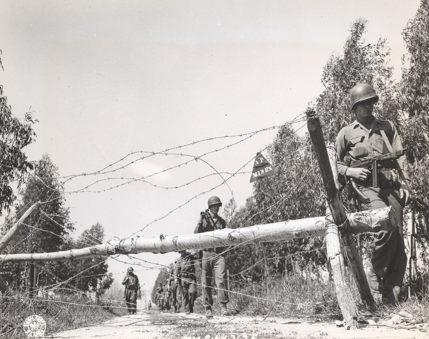 United States Army soldiers patrol near the Gustav Line in Italy during the Battle of Monte Cassino, carefully moving past barbed wire defenses. The closest soldier is armed with a Thompson submachine gun, emphasizing the close-quarters combat often encountered in this rugged terrain. The scene captures the tense atmosphere as troops advance cautiously under the constant threat of German counterattacks. Barbed wire and rough terrain reflect the extensive defensive measures employed along the Gustav Line, a key obstacle in the Italian Campaign. The image illustrates the challenges faced by Allied forces as they worked to break through German fortifications. It highlights the determination and bravery of the soldiers during this pivotal battle.