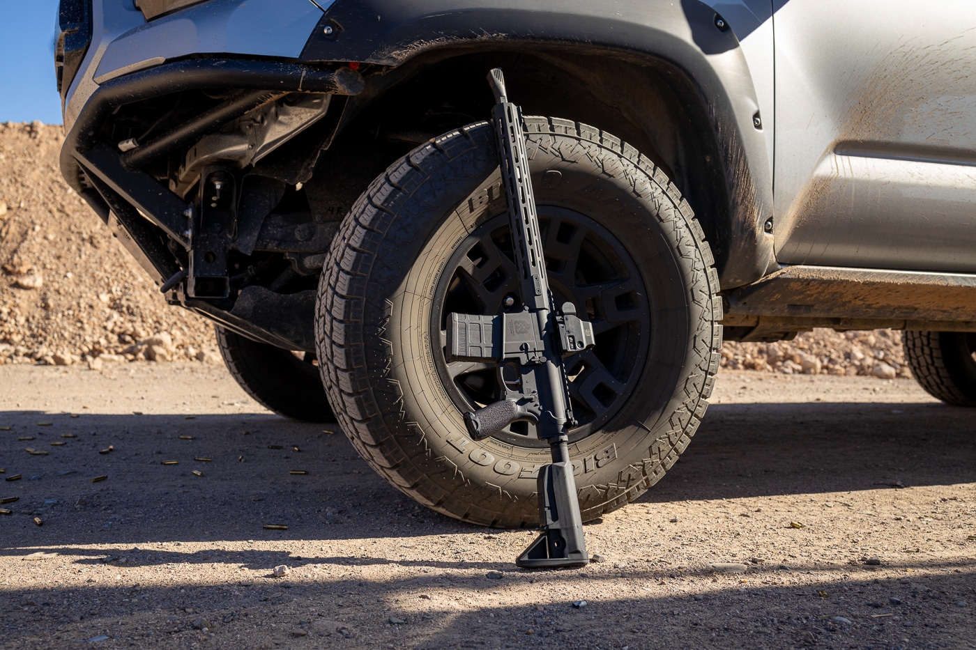 A Springfield Armory SAINT Victor rifle is propped against the tire of a truck at an outdoor shooting range. The rifle, chambered in .308, is equipped with the Walker NERO 762 muzzle brake, which features its distinctive angular design. The truck's tire and rim provide a sturdy background, hinting at the author's practical approach to testing. The firearm’s matte black finish gleams subtly in the natural light, blending seamlessly with the rugged outdoor environment. Dry grass and scattered dirt indicate the authentic, real-world conditions of the range. The rifle is positioned with care, showcasing the attached muzzle brake and overall setup. This photo emphasizes the balance between advanced technology and field-tested durability. The scene reflects the preparation for a live shooting session to evaluate performance.