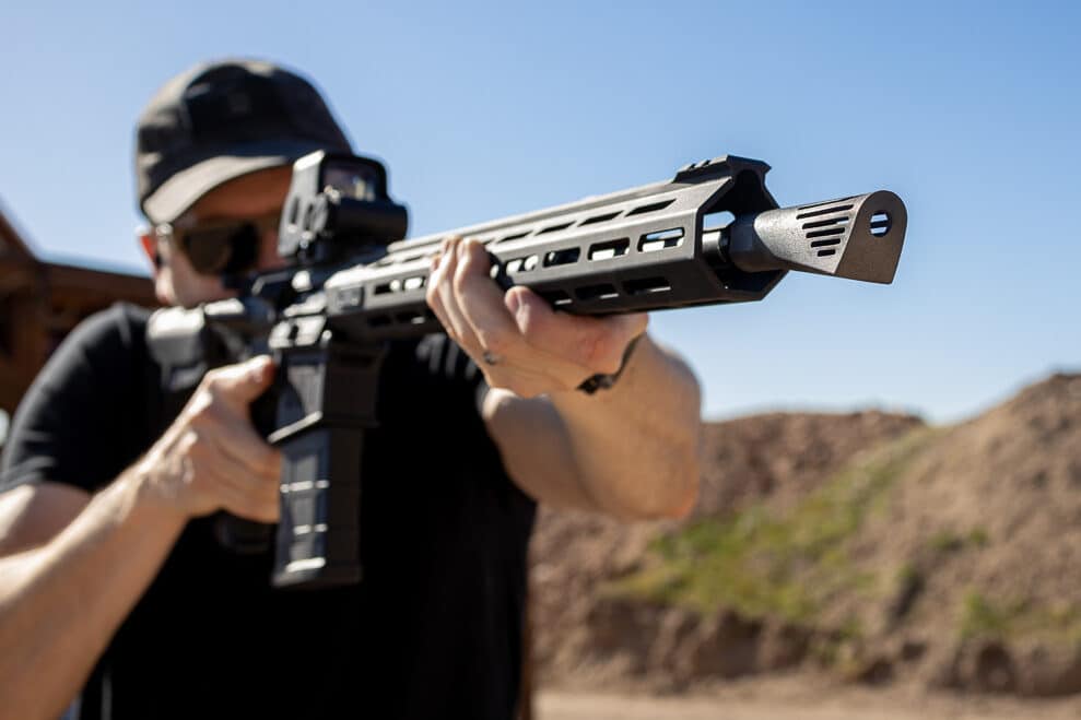 A shooter is testing a Springfield Armory SAINT Victor .308 rifle equipped with the Walker NERO 762 muzzle brake at an outdoor range. The rifle's matte black finish contrasts with the distinct, sharp angles of the NERO muzzle brake at the barrel's end. The shooter maintains a stable stance, with the rifle securely pressed into the shoulder, aiming toward a distant target. The image highlights the innovative engineering of the NERO, designed to neutralize recoil and eliminate muzzle climb. The background consists of open greenery and clear skies, creating a serene yet focused shooting environment. The photograph captures the practical application of cutting-edge firearm technology. The shooter’s stance and expression suggest confidence in the rifle’s performance, supported by the advanced AR10 muzzle brake.