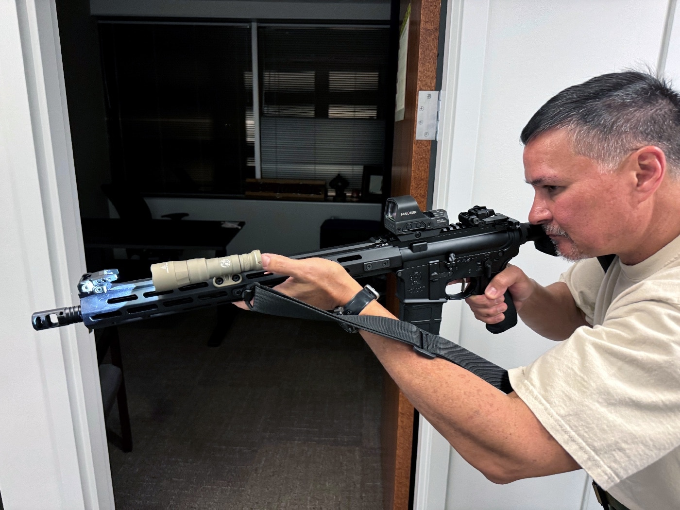 The author navigates an office building during a training exercise, demonstrating how to activate the SureFire Mini Scout Pro flashlight mounted on a Springfield Armory SAINT rifle. The tailcap pressure switch is easily accessed with the thumb, providing quick illumination when needed. The flashlight’s 500-lumen output ensures clear visibility in the dimly lit office, aiding in situational awareness and target identification. The light’s Hybrid Beam Pattern offers both a focused beam for precision and a peripheral spill for a broader view. This scene illustrates the practicality of the flashlight for tactical applications in confined and structured environments.