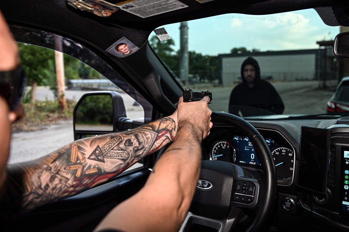 A man inside his vehicle takes careful aim with a Springfield Armory Echelon pistol, pointing through the windshield at an advancing carjacker. The windshield remains intact but shows signs of dirt and glare, emphasizing the confined and challenging conditions of aiming through glass. Outside the vehicle, the carjacker is partially visible, dressed in dark clothing, and moving aggressively toward the truck. The driver maintains a composed demeanor, demonstrating focused training and control in a life-threatening scenario. The truck's interior is minimal but functional, showcasing the practical setting of the encounter. The scene captures the tension and high stakes of defending oneself while confined in a vehicle. It highlights the importance of tactical training for real-world self-defense situations.