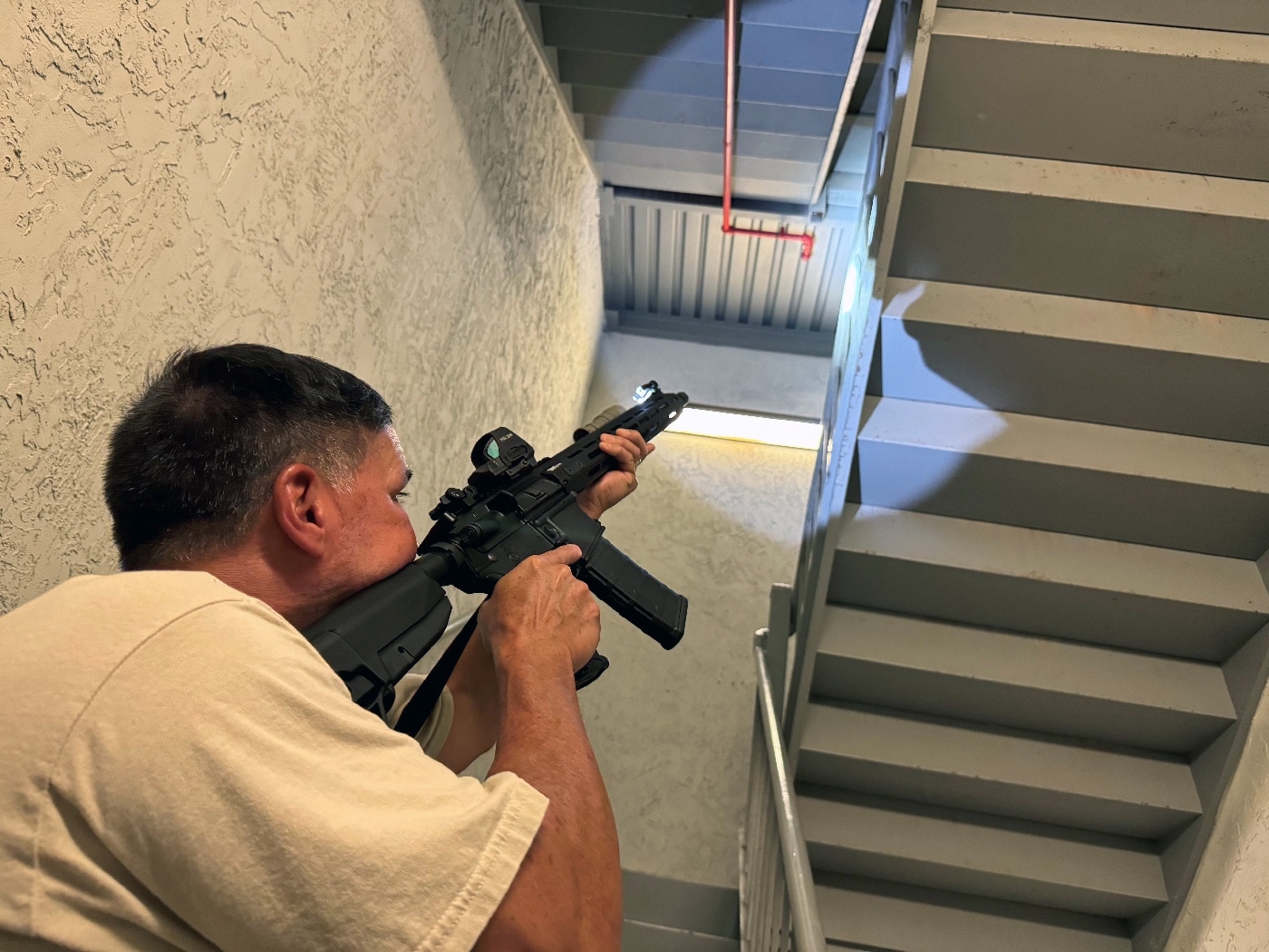 The author participates in a police training exercise, ascending a stairwell with his patrol rifle equipped with a SureFire Mini Scout Pro flashlight. The flashlight’s 500-lumen beam cuts through the shadows, illuminating dark corners and improving visibility in the confined stairwell. Positioned on the M-LOK rail of the rifle, the light allows for seamless activation via the tailcap switch. This setup ensures the author can navigate the stairwell safely and identify potential threats effectively. The scene demonstrates the importance of reliable illumination tools for law enforcement and tactical scenarios.