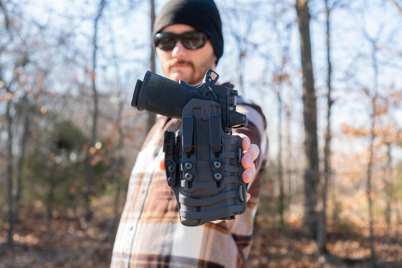 The author is holding the PHLster Floodlight2 holster, emphasizing its lightweight and ergonomic design for concealed carry. The holster’s wave-pattern ComfortScape texture is clearly visible, designed to enhance airflow and reduce pressure during wear. Its durable Kydex construction provides a secure fit for light-bearing pistols, indexing retention off the weapon-mounted light. The open-ended design supports advanced firearm features like threaded barrels and red dot optics, adding to its versatility. The attached red bungee cord is seen, which adjusts the retention for compatibility with a wide range of pistol frames. The holster is designed for inside-the-waistband carry, with sturdy clips ensuring it stays securely attached. This image highlights the Floodlight2's innovative approach to comfort and concealability for concealed carry enthusiasts.