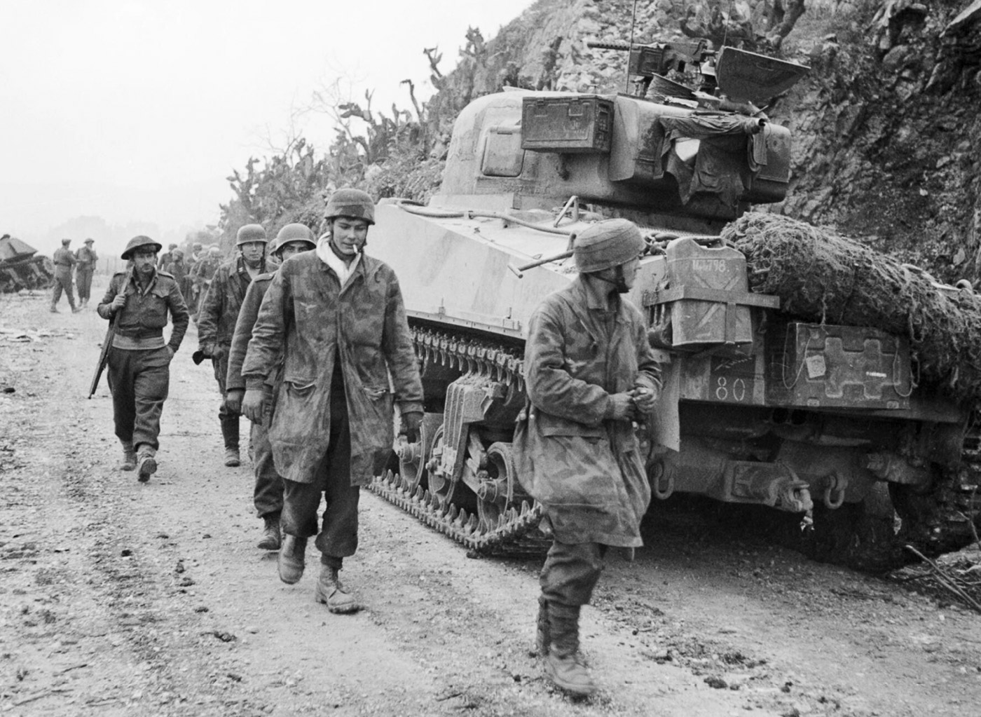 Captured German parachute troops, known as Fallschirmjäger, are seen filing past a Sherman tank of the New Zealand 4th Armoured Brigade on Via Caira in Cassino on 16 March 1944. The paratroopers, part of the German forces defending the Gustav Line, appear subdued as they move under Allied guard following their capture. The Sherman tank in the background underscores the significant role of armored units in the Allied efforts to secure Cassino during the Battle of Monte Cassino. The scene reflects the intense and costly fighting that ultimately led to the German withdrawal. This moment highlights the turning point in the Italian Campaign as Allied forces pushed toward victory.