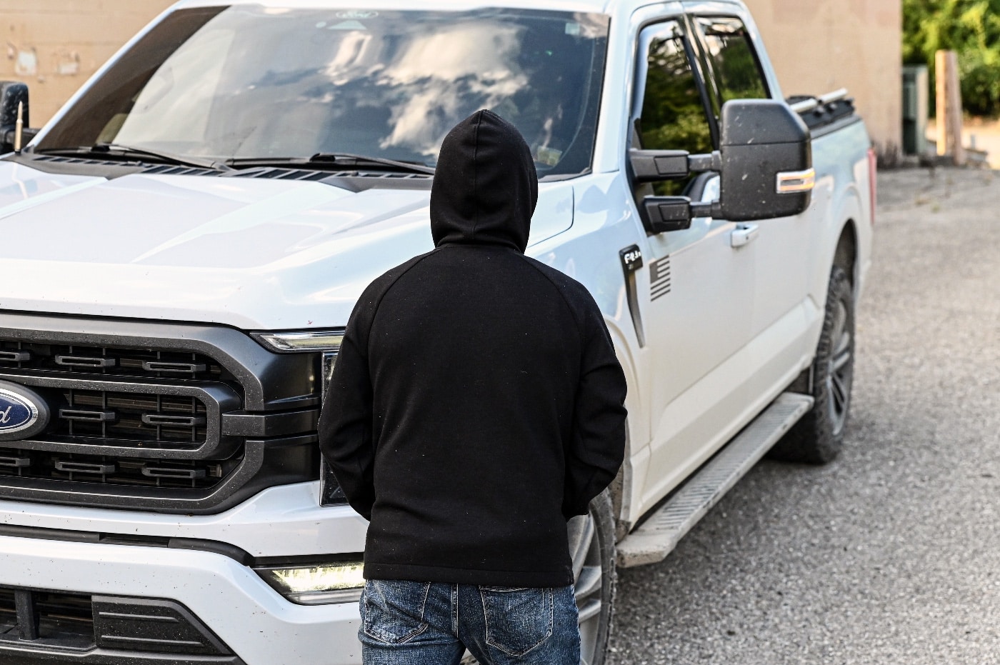 A man dressed in black, wearing a ski mask, approaches a vehicle in a threatening manner, his posture indicating intent to carjack. The driver, inside the truck, is partially visible, gripping his firearm as he assesses the rapidly escalating situation. The urban setting, with traffic lights and vehicles surrounding the intersection, suggests a populated area where such incidents are least expected. The would-be attacker’s hand positioned behind his back adds to the tension, implying potential concealed danger. The scene captures the pivotal moment when self-defense training and awareness become critical for survival. The boxed-in vehicle highlights the challenges faced by the driver, emphasizing the importance of tactical decision-making. The overall composition of the image reflects the seriousness and urgency of real-world personal defense scenarios.