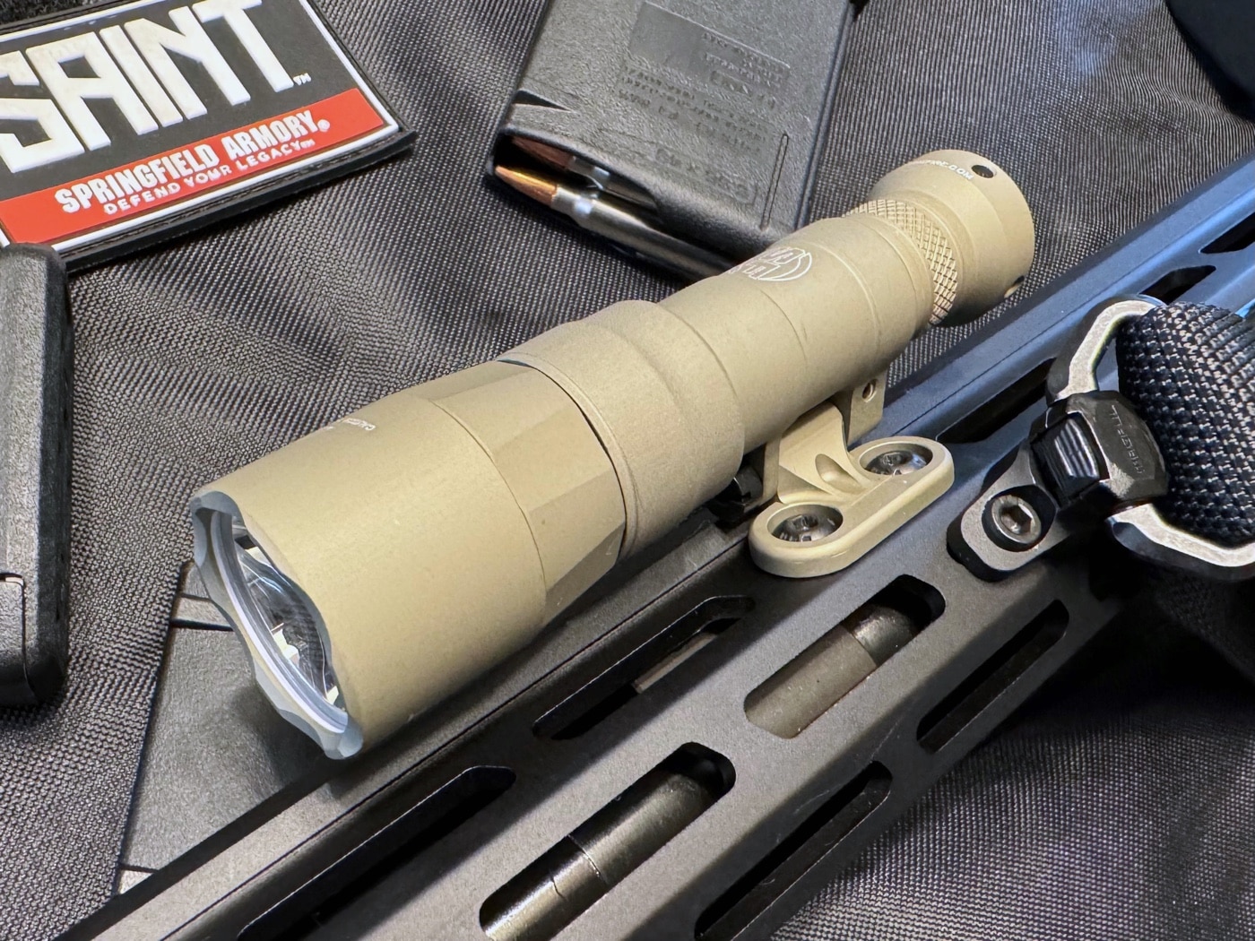 A close-up of the SureFire Mini Scout Pro flashlight in Flat Dark Earth (FDE) color mounted to the M-LOK rail of a Springfield Armory SAINT rifle. The flashlight's durable aluminum body features a hard coat anodized finish for enhanced protection against wear. The low-profile mount holds the light securely along the rifle's foregrip, providing optimal positioning for tactical use. The earthy tones of the FDE finish blend well with the rifle’s design, reflecting both functionality and aesthetics. This image highlights the flashlight’s compact yet robust construction, ideal for professional and self-defense applications.