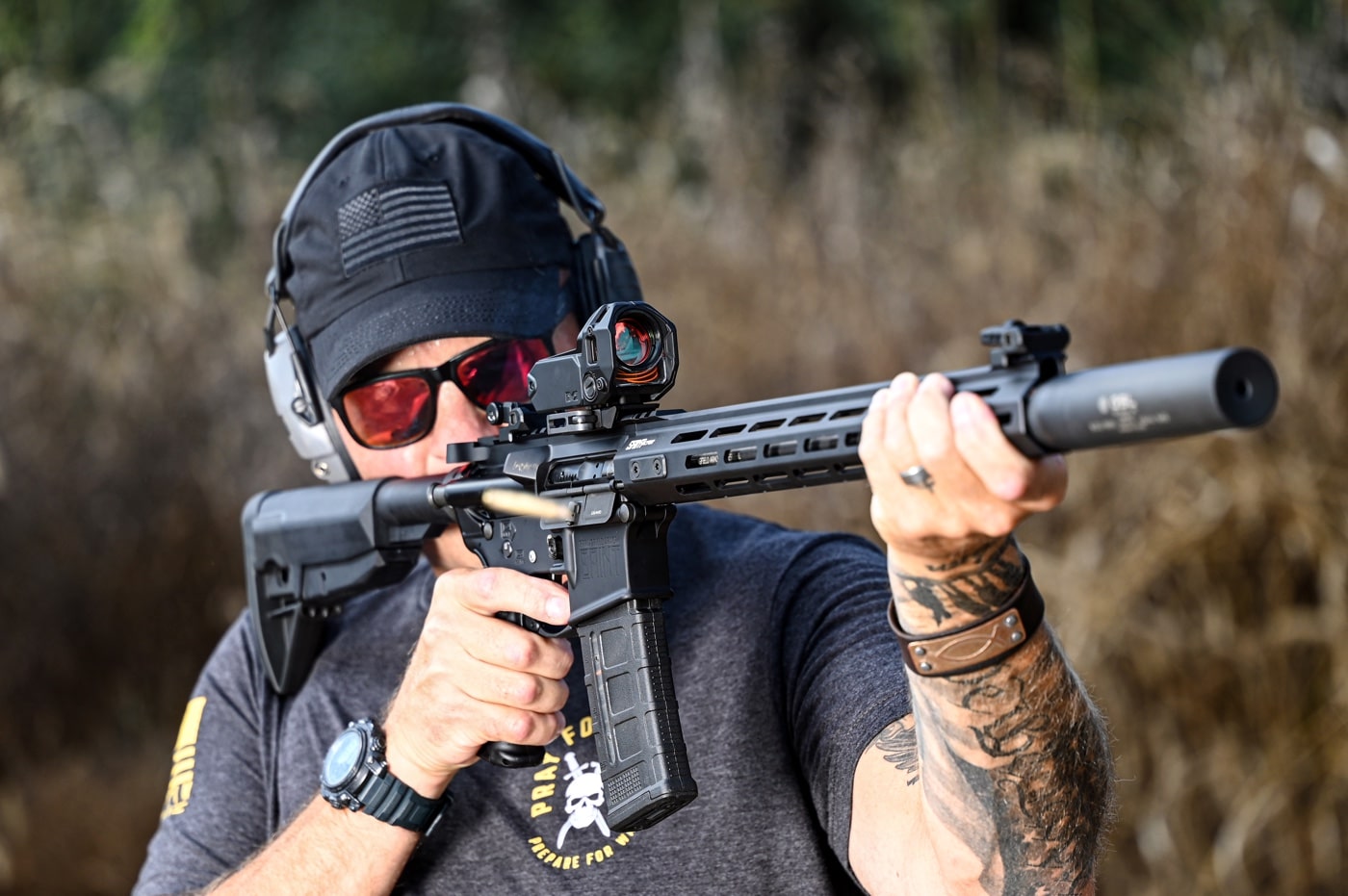 The author is captured shooting a Springfield Armory SAINT rifle outfitted with a Meprolight M22 red dot sight and a suppressor. The suppressor extends the rifle's barrel, demonstrating a focus on reduced noise and improved performance. The M22 optic sits securely on the Picatinny rail, providing a clear reticle powered by fiber optics and tritium. The setup showcases a balance of advanced firearm accessories designed for precision and reliability. The scene emphasizes the practical use of the rifle and optic combination in a dynamic shooting environment.