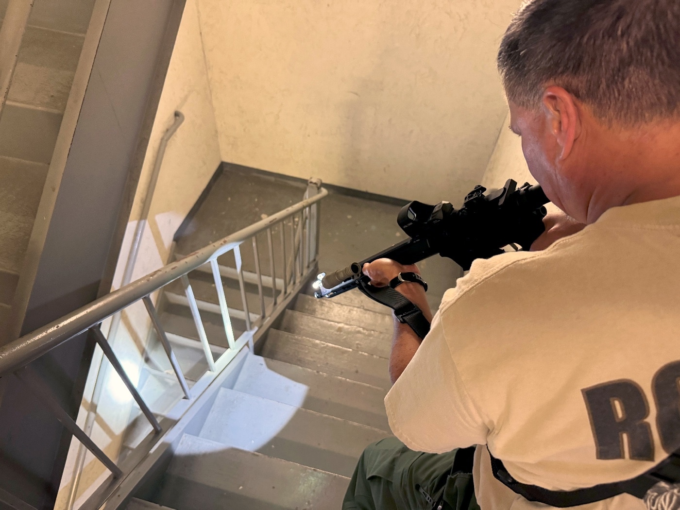 The author tactically clears a stairwell using a Springfield Armory SAINT rifle mounted with the SureFire Mini Scout Pro flashlight. The flashlight emits a 500-lumen beam, illuminating the stairwell with a clear and focused Hybrid Beam Pattern. Its peripheral light spill ensures situational awareness in tight quarters, while its ergonomic design allows intuitive activation via the tailcap switch. The scene illustrates the flashlight's practical application in self-defense or law enforcement scenarios. The overall setup highlights the importance of proper equipment for navigating challenging indoor environments.